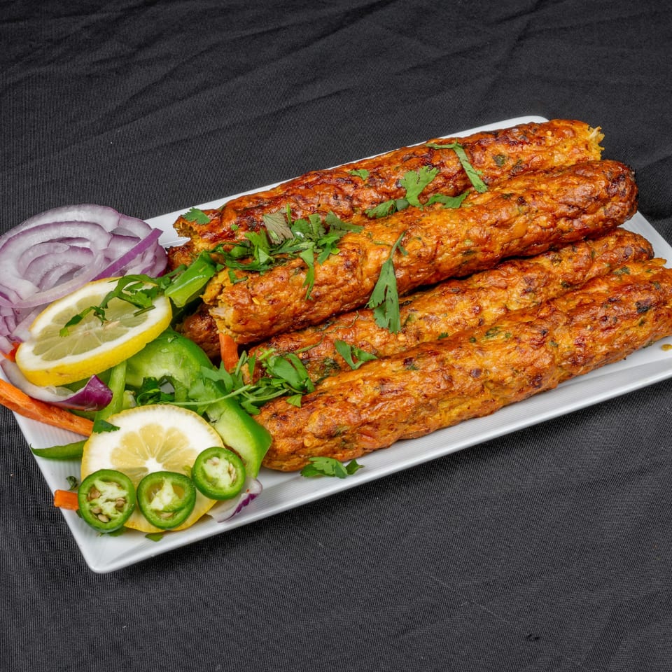 Indian seekh kebabs garnished with cilantro, lemon, red onion, and green peppers on a white plate.