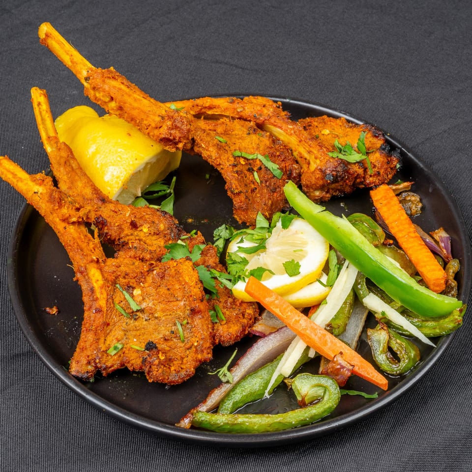 Indian lamb chops with lemon wedges, green peppers, and carrots on a black plate.