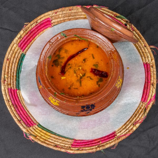 Indian dal in a rustic clay pot with a lid, garnished with red chilies, on a woven mat.